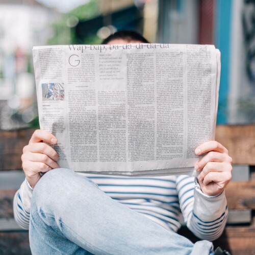 Eine Person sitzt auf einer Palettenbank und liest eine Zeitung. Die Zeitung ist so hoch aufgerichtet, dass man nur noch die Stirn der Person sieht