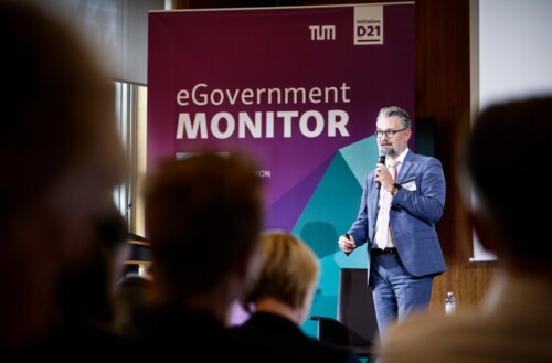 André Göbel spricht mit einem Mikrofon in der Hand auf der Bühne des GovTalk. Im Hintergrund ein großes Roll-up zum eGovernment MONITOR. Im Vordergrund verschwommen die Hinterköpfe von inigen Menschen im Publikum