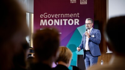 André Göbel spricht mit einem Mikrofon in der Hand auf der Bühne des GovTalk. Im Hintergrund ein großes Roll-up zum eGovernment MONITOR. Im Vordergrund verschwommen die Hinterköpfe von inigen Menschen im Publikum