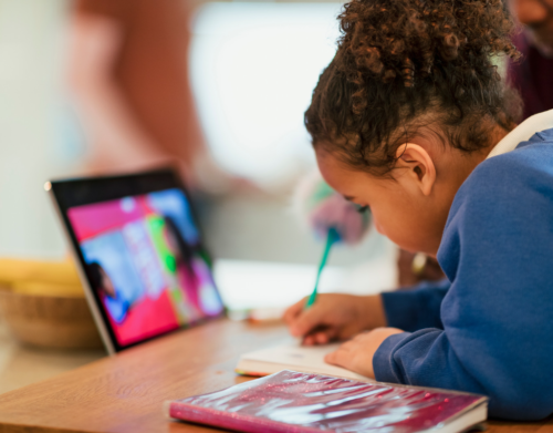 Ein Kind sitzt vor einem Tablet, auf dem ein Video läuft, und schreibt dabei etwas in ein Schulheft.