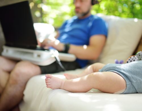 Im Hintergrund sitzt eine Person an einem Laptop auf der Couch. Im Vordergrund sieht man die beine eines Babys, das ebenfalls auf der Couch liegt.