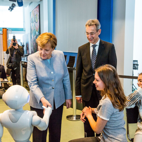 Angela Merkel schüttelt einem Peppa-Roboter die Hand. Hannes Schwaderer und drei Schülerinnen schauen ihr dabei zu.