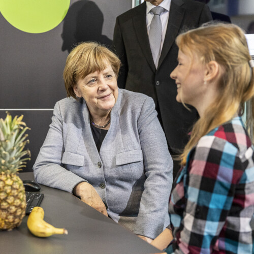 Angela Merkel und eine Schülerin unterhalten sich am Stand von Deloitte. Vor ihnen auf dem Tisch liegen eine Melone, eine Ananas und eine Banane.