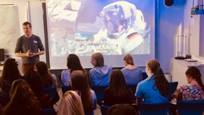 Schülerinnen sitzen vor einer Leinwand, auf der ein Astronaut zu sehen ist