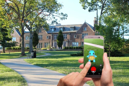 Ein Handy, das von zwei Händen gehalten wird, mit der App Pokémon Go mit dem Pokémon "Shiggy" auf dem Display vor einem großen Landhaus und einer grünen Wiese.
