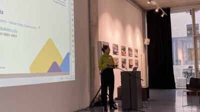 Eva Schmitz steht an einem Rednerpult vor einer an die Wand geworfenen Powerpoint-Präsentation