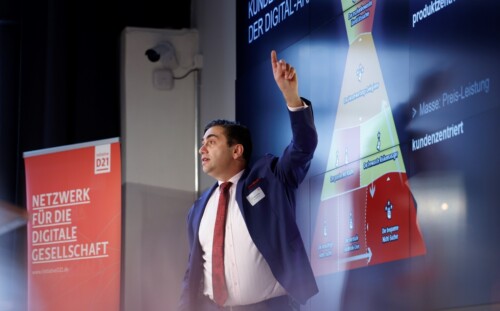 Sven Gábor Jánszky steht vor seiner Präsentation, die hinter ihm auf einem riesigen Bildschirm gezeigt wird, und zeigt dem Publikum mit dem Finger etwas darauf.