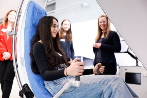 Eine Schülerin sitz auf einem Sitz und greift mit ihren Händen zwei Hebel, die seitlich an dem Sitz montiert sind und ihre Vitaldaten messen.