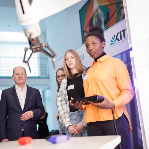 Eine Schülerin bedient einen Roboterarm am Stand vom KIT (Karlsruher Institut für Technologie). Kanzler Scholz und D21-Präsident Schwaderer schauen zu.