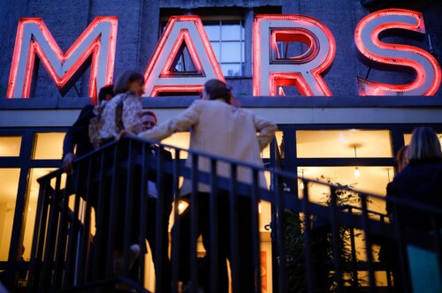 Die Leuchtschrift des MARS-Gartens leuchtet rot, davor auf der Treppe unterhalten sich Menschen.
