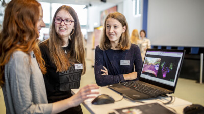 Eine weibliche Person erklärt zwei Schülerinnen etwas an einem Laptop.
