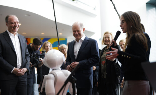 Der Bundeskanzler, Marc Reinhardt und einige Schülerinnen interagieren mit einem weißen Roboter. Im Hintergrund Medienvertreter*innen.