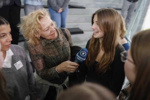 Eine Schülerin steht im Kreis anderer Schülerinnen und wird von einer Journalistin mit einem Mikrofon in der Hand interviewt.