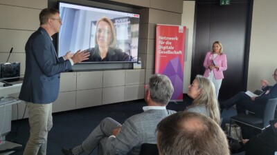 Ernst Bürger und Cornelia Gottbehüt stehen vor einem großen Screen, auf dem Laura Dornheim eingeblendet wird. Im Vordergrund das Publikum, das sie anschaut.