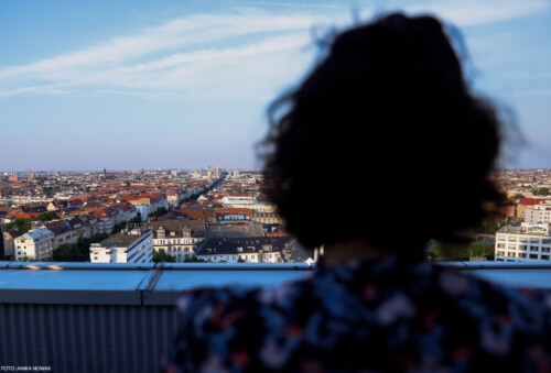 Blick über Berlin von der Dachterrasse, dazu der in Nahaufnahme verschwommene Hinterkopf einer Person, die in die Ferne schaut