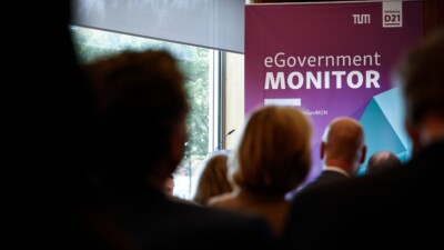 Das Publikum von hinten fotografitert. Im Hintergrund ist scharf ein eGovernment MONITOR-Banner zu erkennen