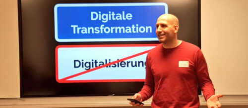 Ein Foto von Dejan Mihajlovic, wie er vor einem großen LCD-Bildschirm während seines Impuls spricht. Der Bildschirm zeigt zwei farbige Kästen mit Text: Im oberen Kasten steht "Digitale Transformation", während der untere Kasten durchgestrichen ist und den Begriff: "Digitalisierung" enthält.
