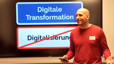 Ein Foto von Dejan Mihajlovic, wie er vor einem großen LCD-Bildschirm während seines Impuls spricht. Der Bildschirm zeigt zwei farbige Kästen mit Text: Im oberen Kasten steht "Digitale Transformation", während der untere Kasten durchgestrichen ist und den Begriff: "Digitalisierung" enthält.